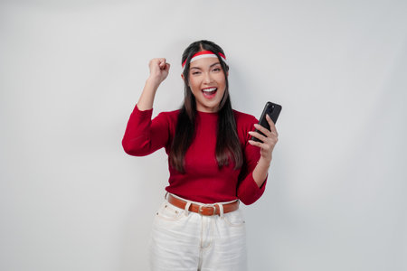 Excited Asian woman in a red shirt holding a cell phone and while raising fist to celebrate victory or a happy moment.の写真素材