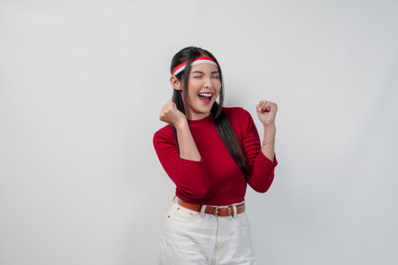 Excited young Asian woman in a red shirt and white pants is raising her hands in the air with happy successful expression.の写真素材