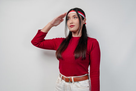Beautiful young Asian woman in a red shirt and white pants doing salute pose to country flag. Indonesia Independence Day concept.の写真素材