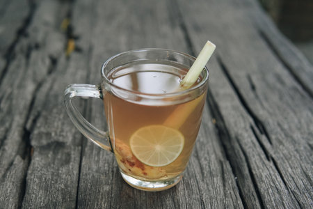 A glass of tea with a slice of lime and lemon grass in it sits on a wooden table with nature background. Concept of relaxation and enjoyment.の写真素材
