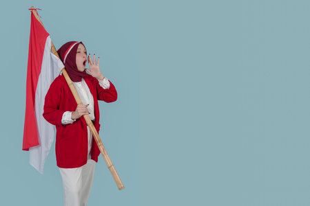 Beautiful Indonesian woman wearing hijab holding a flag and yelling with hands and mouth wide...