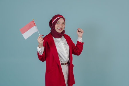 Attractive Indonesian woman wearing a red jacket and hijab holding a mini flag while raising fist gesture. Indonesian Independence Day concept.の写真素材