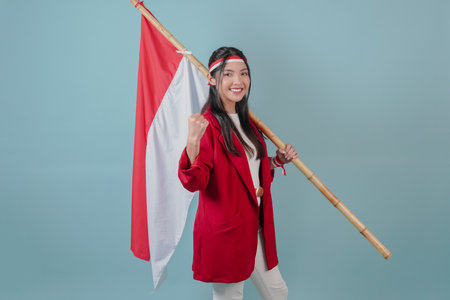 Smiling Indonesian woman wearing a red jacket and white pants is holding a flag while raising fist gesture. Indonesian Independence Day concept.の写真素材