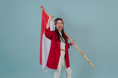 Smiling Indonesian woman wearing a red jacket and white pants is holding a flag while raising fist gesture. Indonesian Independence Day concept.の写真素材