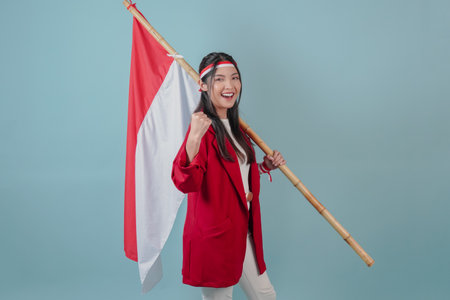 Smiling Indonesian woman wearing a red jacket and white pants is holding a flag while raising fist gesture. Indonesian Independence Day concept.の写真素材