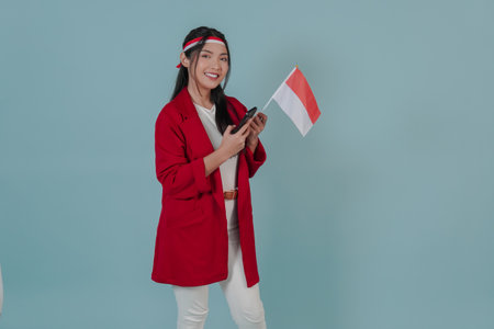 Smiling young Indonesian woman wearing a red jacket and holding a mini flag while using smartphone to browse internet. Indonesia Independence Day concept.の写真素材