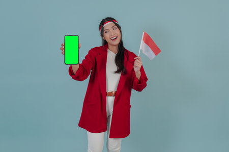 Cute young Indonesian woman holding a mini country flag while showing copy space on her phone screen. Indonesia Independence Day concept.の写真素材