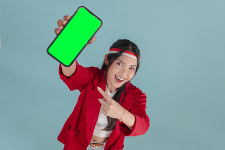 Beautiful young Indonesian woman smiling while pointing to the copy space on her phone screen, isolated by blue background. Indonesia Independence Day concept.の写真素材