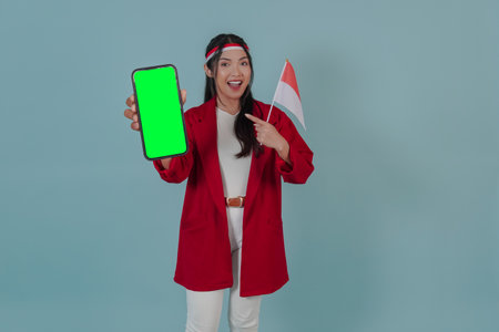 Joyful Indonesian woman holding a mini country flag while pointing to the copy space on her phone screen. Indonesia Independence Day concept.の写真素材
