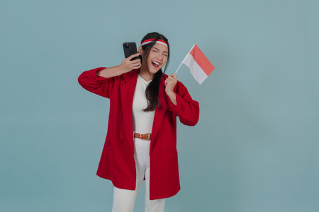 Excited young Indonesian woman holding a mini flag and smartphone with a happy successful expression. Indonesia Independence Day concept.の写真素材
