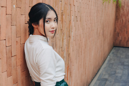 Beautiful young Asian woman in a green skirt stands in front of a brick wall. She is wearing a white shirt.の写真素材