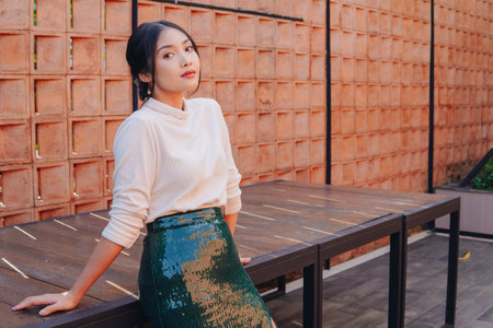 Beautiful young Asian woman in a green sequined skirt stands in front of a brick wall. She is wearing a white shirt and heels.の写真素材