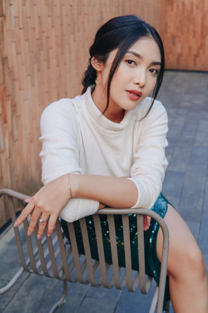 Beautiful young Asian woman in a green sequined skirt sits on a chair. She is wearing a white shirt.の写真素材