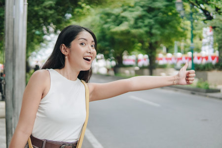 Beautiful young Asian woman wearing a white shirt is standing on the side of the road, giving a thumbs up to a driver. Holiday and vacation concept.の写真素材