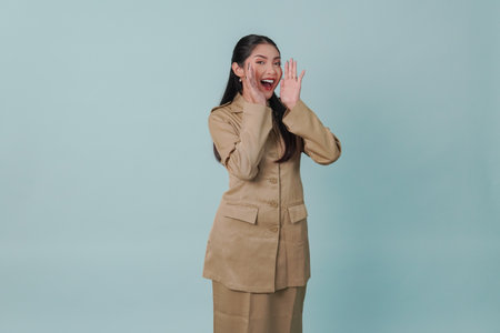 Joyful Indonesian government worker woman is shouting or yelling isolated by a blue background. Civil servant concept.の写真素材