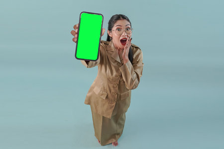 Amazed government worker woman wearing khaki uniform and glasses showing copy space on her phone to the camera on top. PNS civil servant concept.の写真素材