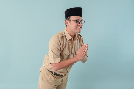 Polite Indonesian government worker man doing greeting gesture while smiling, isolated by blue background. PNS civil servant concept.の写真素材