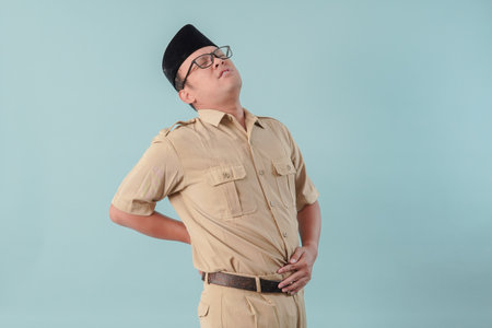 Tired Indonesian government worker man wearing khaki uniform suffering from pain, muscle spasm on his back. PNS civil servant concept.の写真素材