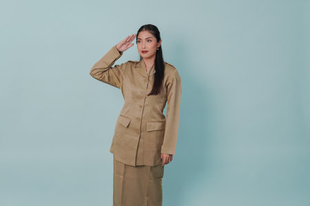 Confident Indonesian government worker wearing a khaki uniform doing salute pose, isolated by blue background. PNS civil servant concept.の写真素材