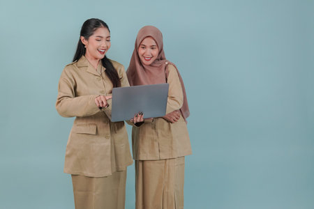 Two cheerful Indonesian government worker women wearing khaki uniform looking at the laptop they hold while smiling. PNS and civil servant concept.の写真素材