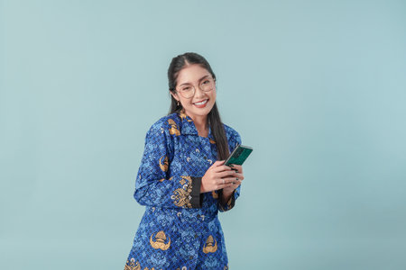 Excited government worker woman wearing blue batik uniform and glasses holding a smartphone. PNS civil servant concept.の写真素材