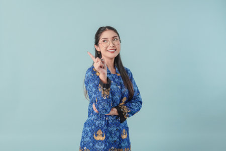 Cheerful government worker woman in blue batik uniform and glasses pointing to the copy space behind her with a smile. PNS civil servant concept.の写真素材
