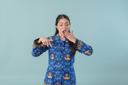 Government worker woman wearing blue batik uniform and glasses pointing to the copy space below with surprised look. PNS civil servant concept.の写真素材