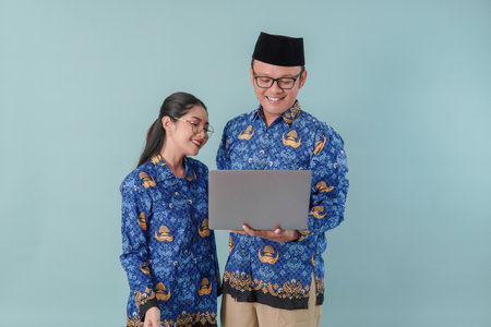 Two smiling government workers in blue batik uniform and glasses looking on the laptop. PNS civil servant concept.の写真素材