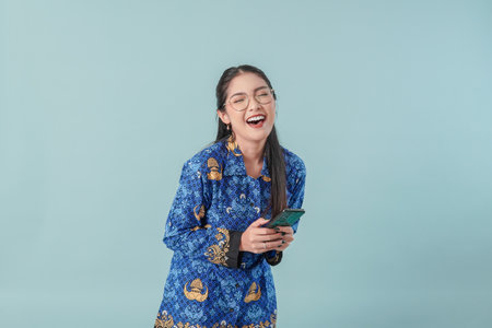 Excited government worker woman wearing blue batik uniform and glasses holding a smartphone. PNS civil servant concept.の写真素材
