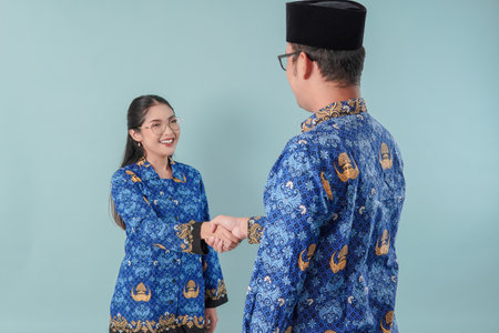 Calm Indonesian government workers wearing blue batik uniform doing handshake gesture, isolated by blue background. PNS civil servant concept.の写真素材
