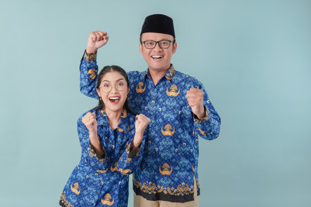 Two excited Indonesian government workers wearing blue batik uniform raising fist with a happy successful expression. PNS and civil servant concept.の写真素材