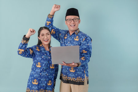 Two excited Indonesian government workers wearing blue batik uniform holding laptop with a happy successful expression. PNS and civil servant concept.の写真素材