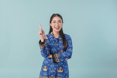 Happy government worker woman wearing blue batik uniform and glasses pointing to the copy space upward. PNS civil servant concept.の写真素材
