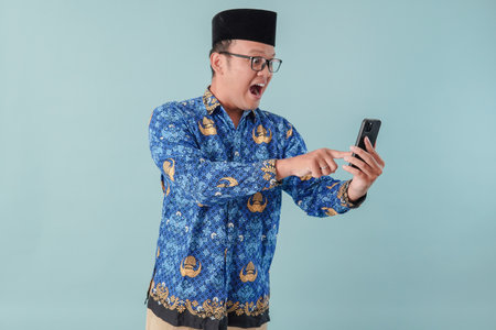 Happy government worker man wearing blue batik uniform doing video call while pointing to his smartphone. PNS civil servant concept.の写真素材