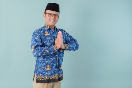 Cheerful Indonesian government worker man wearing blue batik uniform doing greeting gesture while...