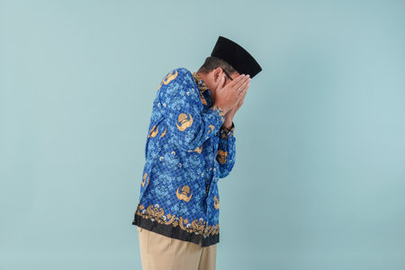 Scared government worker man wearing blue batik uniform with face covered by hands frightened and...