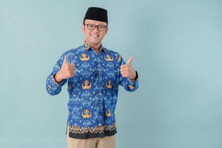 Handsome Indonesian government worker man wearing blue batik uniform smiling and giving thumb up gesture. PNS civil servant concept.の写真素材