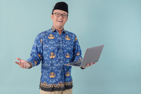 Excited government worker man wearing blue batik uniform holding laptop and looking to the camera....
