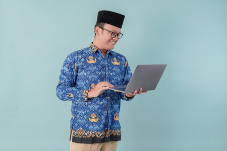 Smiling face government worker man wearing blue batik uniform watching streaming video or news on laptop. PNS and civil servant concept.の写真素材