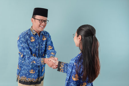 Calm Indonesian government workers wearing blue batik uniform doing handshake gesture, isolated by blue background. PNS civil servant concept.の写真素材