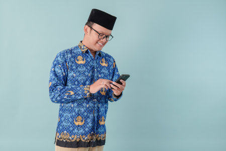 Handsome government worker man wearing blue batik uniform holding smartphone while smiling. PNS and civil servant concept.の写真素材