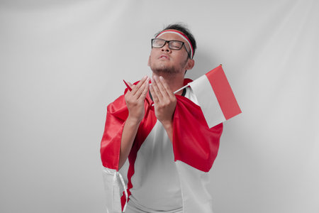 Serious Indonesian man wearing jersey with flag draped over shoulder doing praying gesture while watching sport match. Indonesia Independence Day concept.の写真素材
