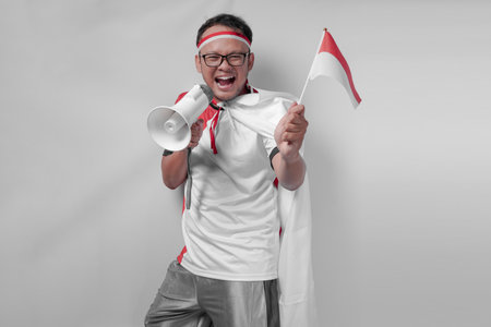 Happy young Indonesian man wearing jersey is holding a mini country flag while shouting at loudspeaker. Indonesia Independence Day and sport concept.の写真素材
