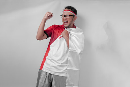 Energetic young Asian man wearing jersey with flag draped over shoulder raising hand in the air with happy expression. Indonesia Independence Day and sport concept.の写真素材