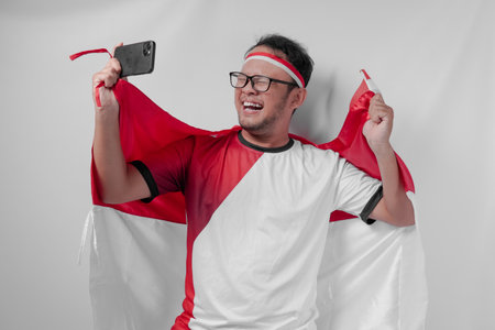 Joyful Indonesian man holding flag over his back and smartphone while raising fist to celebrate something. Indonesia Independence Day and sport concept.の写真素材