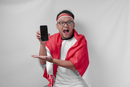 Smiling Indonesian man wearing jersey presenting copy space on his phone over isolated white background. Indonesia Independence Day and sport concept.の写真素材