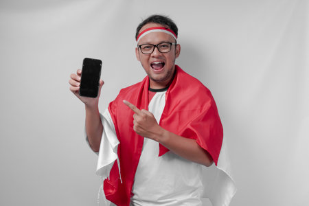 Happy Indonesian man wearing jersey pointing to his phone over isolated white background. Indonesia Independence Day and sport concept.の写真素材