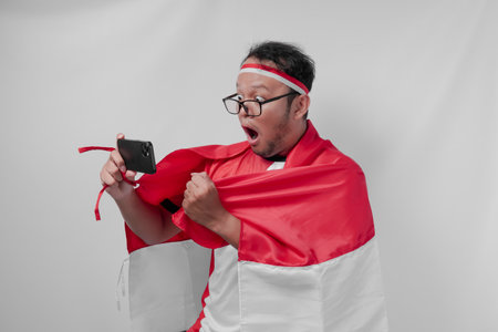 Joyful Indonesian man holding flag over his back and smartphone while raising fist to celebrate something. Indonesia Independence Day and sport concept.の写真素材
