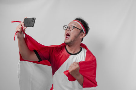 Joyful Indonesian man holding flag over his back and smartphone while raising fist to celebrate something. Indonesia Independence Day and sport concept.の写真素材