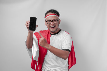 Happy Indonesian man wearing jersey pointing to his phone over isolated white background. Indonesia...
